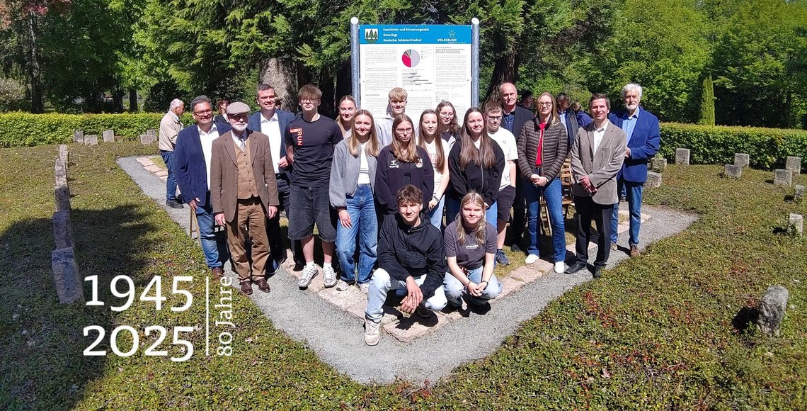 Eine Gruppe Jugendlicher und Erwachsener auf einer Kriegsgräberstätte. Rundherum Grabsteine.Hinter der Gruppe die Geschichts- und Erinnerungstafel.