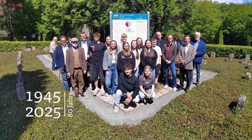 Eine Gruppe Jugendlicher und Erwachsener auf einer Kriegsgräberstätte. Rundherum Grabsteine.Hinter der Gruppe die Geschichts- und Erinnerungstafel.