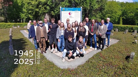 Eine Gruppe Jugendlicher und Erwachsener auf einer Kriegsgräberstätte. Rundherum Grabsteine.Hinter der Gruppe die Geschichts- und Erinnerungstafel.