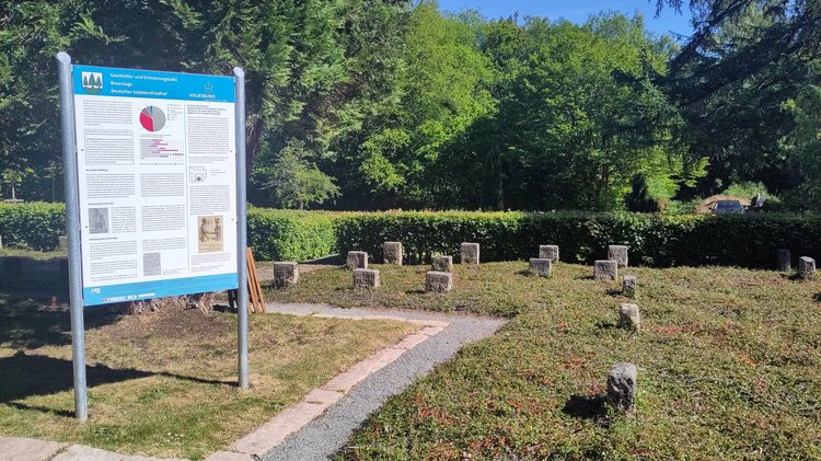 Die Geschichts- und Erinnerungstafel in einem Metallgestell. Darum Grabsteine auf einer Rasenfläche